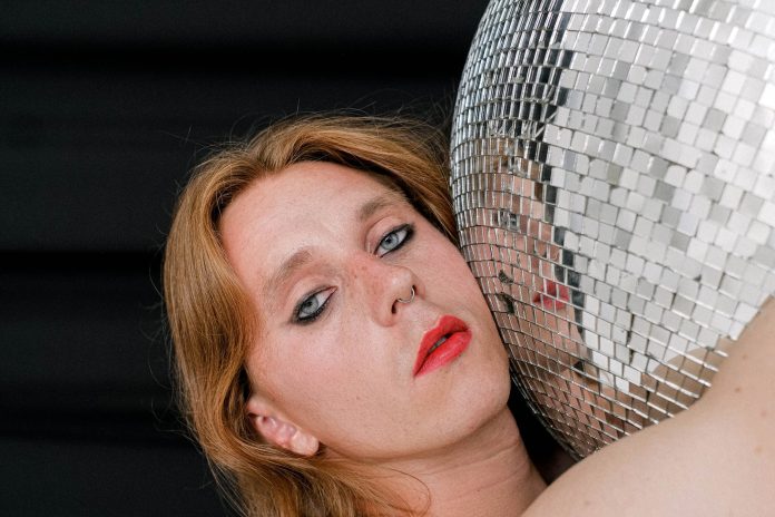 image-16 Pensive young androgynous man with long hair and makeup hugging mirrored disco ball and looking at camera against black background