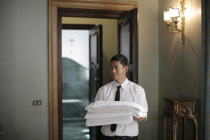 image-73 Young male housekeeper carrying stack of white bed sheets while entering bedroom in hotel