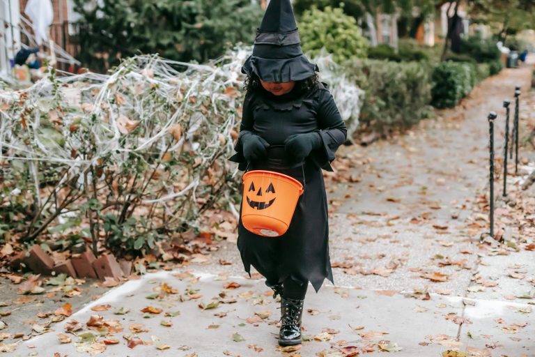 Full body anonymous of little girl in black witch costume for Halloween walking with bucket on street in autumn in daytime