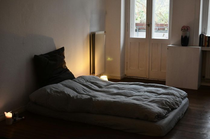 image-110 Comfortable mattress with blanket and cushion placed on wooden floor in dark bedroom with burning candles and flowers in vase