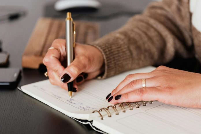 image-16 Crop unrecognizable female with stylish manicure sitting at black desk with keyboard and smartphone and taking notes with silver pen in notepad